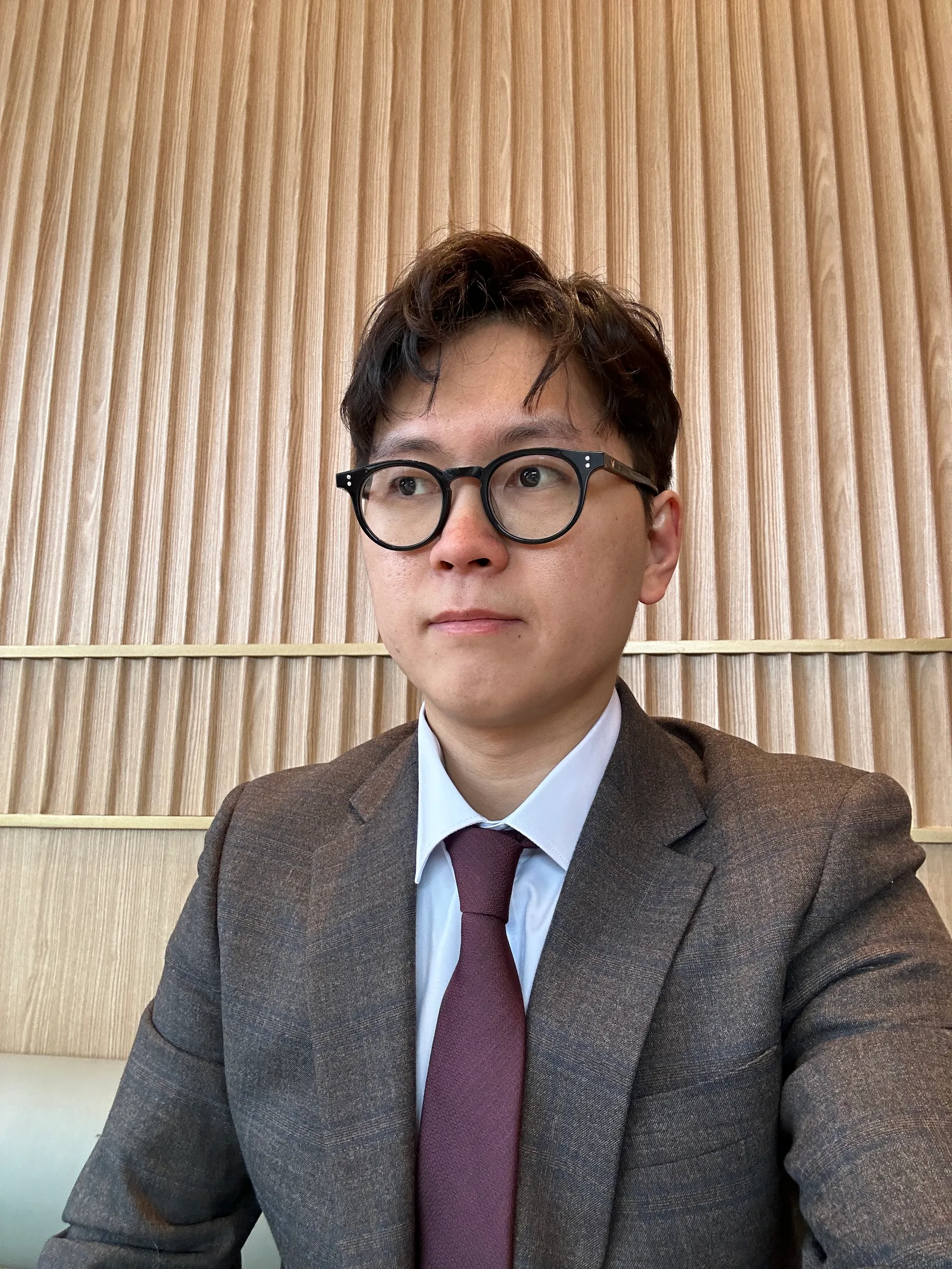 갈색 정장에 흰 셔츠, 자주색 넥타이를 매고 안경을 쓴 남성이 우드패널 벽 앞에 앉아 있다.